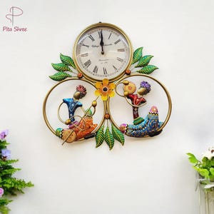 May include: A colorful metal wall clock with a round clock face and two circular frames featuring painted figures. The clock face has Roman numerals and the text "Village Handicrafts" and "Ramnagar, U.P. India".