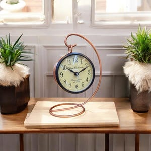 May include: A black and cream clock with a copper stand. The clock face has black Roman numerals and the words "Village Clockworks est 1798 Birmingham" printed on it.
