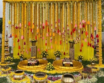 May include: A vibrant yellow and gold floral decoration for a special event. The backdrop features hanging garlands with colorful tassels. Gold pots and decorative stands are arranged on the ground, creating a festive atmosphere.