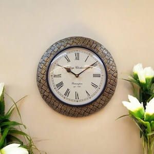 Puede incluir: Un reloj de pared redondo de color dorado con un diseño tejido. La esfera del reloj es blanca con marcas de números romanos negros y agujas negras. La esfera del reloj también incluye el texto "Village Clockwork Birmingham est. 1792".