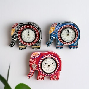 Puede incluir: Tres relojes decorativos con forma de elefante. Los relojes están pintados en negro, azul y rojo con intrincados patrones y esferas circulares. Las esferas tienen números romanos y manecillas negras. Los elefantes tienen diseños detallados en sus cuerpos.