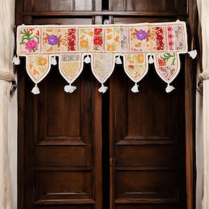Pode incluir: Uma valance de porta de algodão branco com patches florais bordados coloridos e borlas. A valance está pendurada em uma porta de madeira.