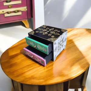 Könnte beinhalten: Eine kleine Holzkiste mit zwei Schubladen. Die Kiste ist schwarz lackiert mit weißen und goldenen Blumendesigns. Die Schubladen sind grün und rosa lackiert mit Blumendesigns.