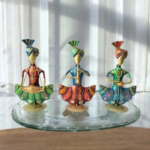 Puede incluir: Tres figuritas de metal de colores de músicos tocando instrumentos. Las figuritas llevan ropa tradicional india y están de pie sobre una bandeja de vidrio. Los músicos tocan un tambor, una flauta y un pandero.