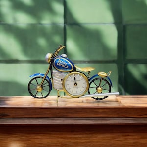 Könnte beinhalten: Eine blaue und goldene Metalluhr in Form eines Royal Enfield Motorrads. Das Zifferblatt der Uhr hat römische Ziffern und einen goldenen Rahmen.