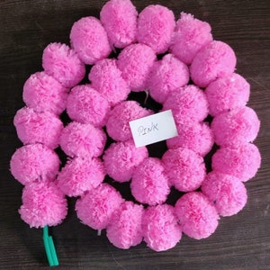 K&ouml;nnte beinhalten: Eine Girlande aus rosa Papierpompons, die mit einer gr&uuml;nen Schnur aneinandergereiht sind. Die Girlande liegt auf einer dunkelbraunen Oberfl&auml;che. Das Wort "PINK" ist auf ein wei&szlig;es Blatt Papier geschrieben.