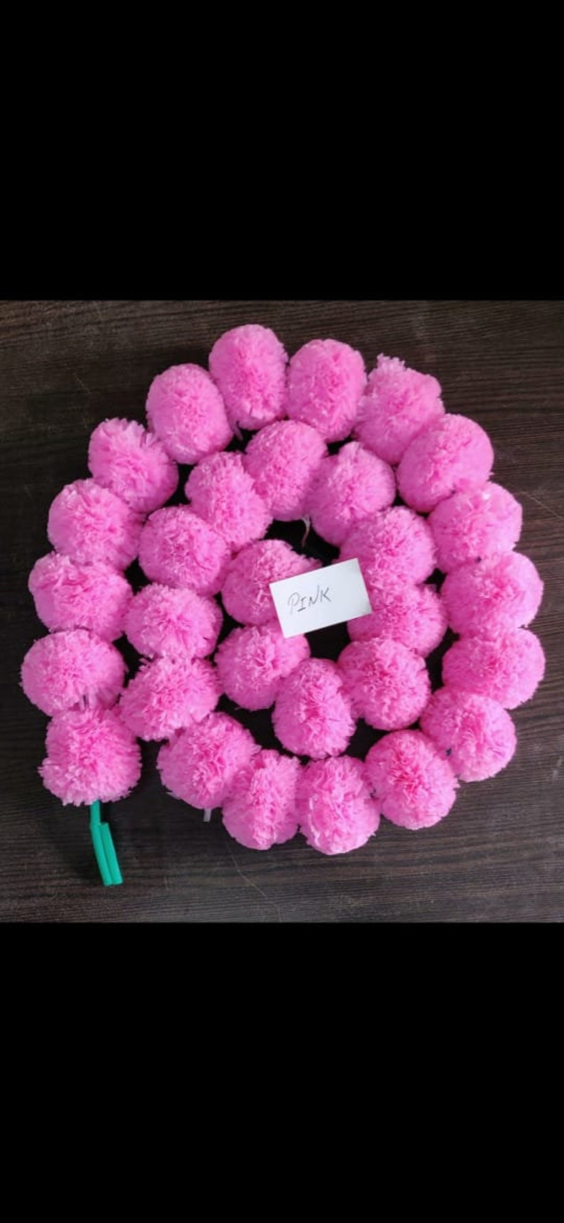 K&ouml;nnte beinhalten: Eine Girlande aus rosa Papierpompons, die mit einer gr&uuml;nen Schnur aneinandergereiht sind. Die Girlande liegt auf einer dunkelbraunen Oberfl&auml;che. Das Wort "PINK" ist auf ein wei&szlig;es Blatt Papier geschrieben.