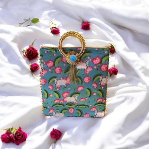 May include: A teal blue clutch bag with a gold ring handle and a tassel. The bag is decorated with a floral pattern of pink lotus flowers and white cows on a teal blue background.