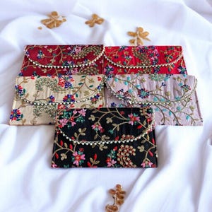 Puede incluir: Una variedad de bolsos de mano bordados en varios colores, incluyendo rojo, beige, rosa y negro. Cada bolso presenta bordados florales y un ribete de cuentas en la solapa. Los bolsos están dispuestos sobre un fondo de tela blanca.