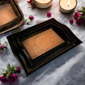 May include: A set of four decorative nesting trays in varying sizes. The trays have a black exterior and a textured, copper-colored interior. The trays are arranged on a white lace tablecloth, with lit candles and rose buds in the background.
