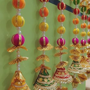 Puede incluir: Una guirnalda decorativa colgante con hilos verticales de esferas de tela multicolor en naranja, amarillo y magenta, intercaladas con perlas blancas y detalles metálicos dorados plisados, terminadas en borlas cónicas adornadas.