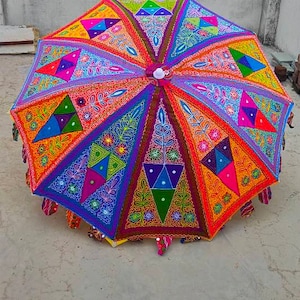 May include: A colourful, multi-patterned, round, fabric umbrella with a fringe border. The umbrella is decorated with embroidered flowers, leaves, and geometric shapes in shades of red, orange, yellow, green, blue, purple, and pink.