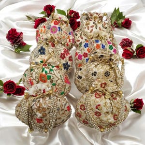 Könnte beinhalten: Ein Set aus acht kleinen, runden, cremefarbenen Kordelbeuteln mit goldenen und perlmuttartigen Verzierungen. Jeder Beutel hat ein anderes florales Stickmuster in verschiedenen Farben, darunter Rot, Rosa, Blau und Grün.