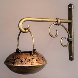 May include: A gold metal wall-mounted candle holder with a decorative scroll design. The holder has a hanging basket with a cut-out pattern.