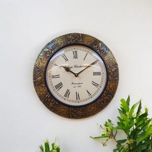 Puede incluir: Un reloj de pared redondo de color dorado con números romanos y una esfera negra. El reloj tiene un diseño detallado y ornamentado alrededor del borde y dice "Village Clockworks Birmingham est 1792".