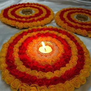 May include: Three round decorative candle holders made of yellow and red fabric roses. The centre holder has a lit tea light candle in a gold metal holder.