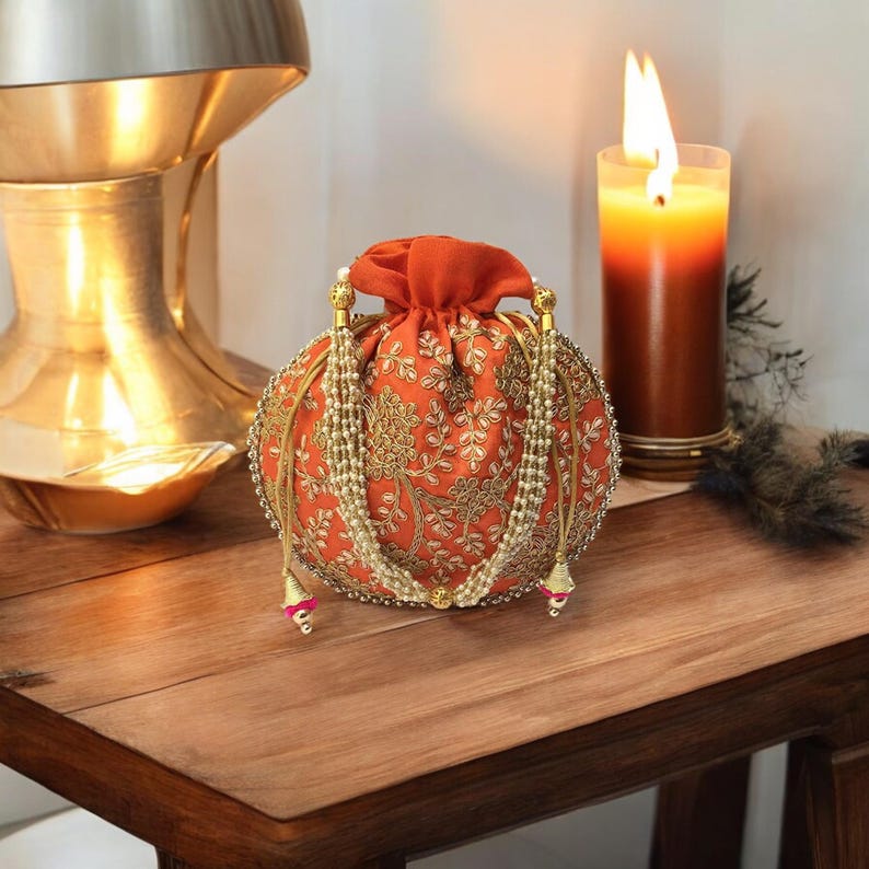 May include: A round orange clutch bag with gold embroidery and pearl accents. The bag has a drawstring closure and is decorated with tassels.