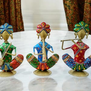 Op de afbeelding: Drie kleurrijke metalen beeldjes van muzikanten die instrumenten bespelen. De beeldjes dragen traditionele Indiase kleding en hebben ingewikkelde details. De muzikant links speelt een snaarinstrument, de muzikant in het midden speelt fluit en de muzikant rechts speelt fluit.