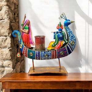 May include: A colourful metal sculpture of a peacock with a couple riding on its back. The peacock is painted in shades of blue, green, and gold, and the couple is painted in bright colours. The sculpture is on a wooden base.