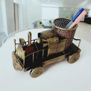 Op de afbeelding: Een decoratieve pennenhouder in de vorm van een vintage trein. De pennenhouder is gemaakt van metaal met een bronzen en gouden afwerking. De trein heeft ingewikkelde details, waaronder decoratieve patronen en juweelaccenten. Twee pennen zijn zichtbaar in de geperforeerde pennenhouder.