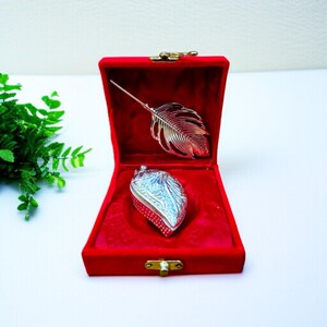 May include: A red velvet jewelry box with a gold clasp, open to reveal a silver feather brooch and a silver leaf-shaped trinket box. The interior of the box is a deep red, creating a striking contrast with the silver items.
