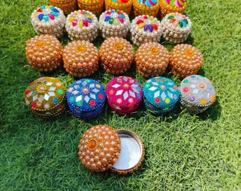 Può includere: Assortimento di piccole scatole rotonde decorative con disegni intricati. Le scatole sono ricoperte di perline e paillettes colorate, e alcune hanno piastrelle a specchio. Le scatole sono disposte a semicerchio su una superficie erbosa verde.
