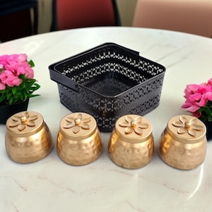 Op de afbeelding: Vier goudkleurige, decoratieve potten met bloemvormige deksels. De potten hebben een textuur en zijn in een rij gerangschikt. Een zwarte, vierkante mand met een decoratief patroon staat achter de potten. Roze bloemen in zwarte potten staan aan weerszijden.