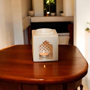 May include: White ceramic oil diffuser with a decorative cutout design and a small candle burning inside. The diffuser is sitting on a wooden table.