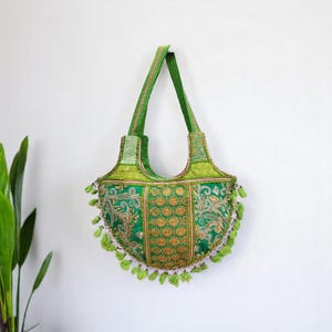 Puede incluir: Un bolso tote verde de patchwork con bordados dorados y borlas verdes. El bolso tiene una sola correa y una forma curva.
