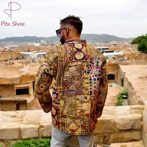 Puede incluir: Un hombre lleva una chaqueta de manga larga multicolor con bordados e incrustaciones intrincadas. La chaqueta está hecha de un mosaico de diferentes telas y presenta una variedad de patrones geométricos. El hombre está de pie frente a una ciudad.