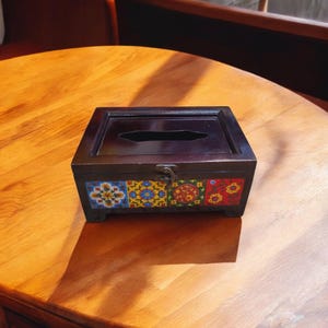 Puede incluir: Un porta pañuelos de madera con un acabado marrón oscuro y diseños de azulejos florales coloridos. La caja tiene una tapa con bisagras y un pequeño pestillo de metal.