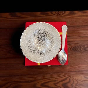 May include: A silver bowl and spoon set with detailed floral designs. The bowl has a decorative edge and rests on a red box. The spoon is positioned beside the bowl. The set is displayed on a dark wooden surface.