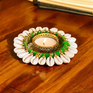 Puede incluir: Un portavelas de conchas blancas con ribete dorado y verde. Una pequeña vela blanca está ardiendo dentro del portavelas.
