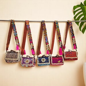 May include: Five colorful embroidered crossbody bags with brown straps and pink tassels hanging on a metal rod. The bags are decorated with beads, shells, and other embellishments.
