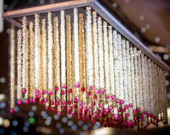 May include: A white floral garland chandelier with pink accents. The garland is hung from a metal frame and is made of white flowers and pink beads.