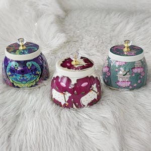 May include: Three decorative ceramic jars with lids. The jars feature colourful designs, including a peacock, cows, and lotus flowers. Each jar has a crystal-like knob on the lid. The jars are set on a fluffy white surface.