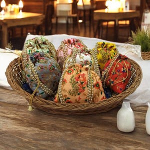 Puede incluir: Una cesta de mimbre llena de bolsas con cordón de colores. Las bolsas presentan estampados florales en tonos rosa, naranja, verde y rojo, con cordones de cuentas. La cesta está sobre una superficie de madera.