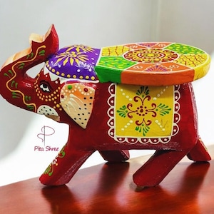 Może przedstawiać: Drewniany stołek w kształcie słonia, ręcznie malowany, z kolorowym, wielobarwnym wzorem. Słoń jest czerwony z żółtymi, zielonymi i fioletowymi akcentami. Stołek jest okrągły i ma płaską górę.