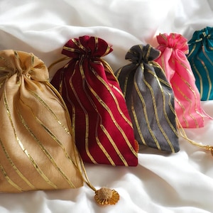 Puede incluir: Una colección de bolsas de regalo con cordón en varios colores, incluyendo dorado, burdeos, gris, rosa y turquesa. Cada bolsa presenta rayas doradas y un cordón a juego con una borla decorativa. Las bolsas están sobre una superficie blanca y sedosa.