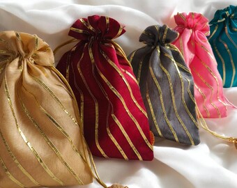 May include: A collection of drawstring gift bags in various colors, including gold, burgundy, gray, pink, and teal. Each bag features gold stripes and a matching drawstring with a decorative tassel. The bags are arranged on a white, silky surface.
