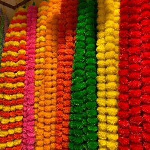 K&ouml;nnte beinhalten: Eine Sammlung bunter k&uuml;nstlicher Blumenketten, darunter Gelb, Orange, Rosa, Gr&uuml;n und Rot. Die Girlanden bestehen aus kleinen, runden Bl&uuml;tenformen und sind zu langen, dekorativen Str&auml;ngen zusammengef&uuml;gt.