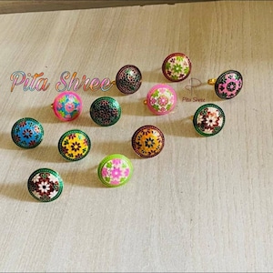 Puede incluir: Surtido de 12 anillos coloridos con intrincados diseños florales. Los anillos están hechos de metal y tienen un acabado de esmalte verde. Los anillos están dispuestos en círculo sobre una superficie de madera. El texto "Pita Shree" es visible en la imagen.