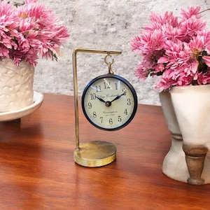 May include: A gold metal stand with a black clock hanging from it. The clock face is white with black numbers and hands. The clock says "Village Clocksmith" and "Birmingham est. 1782".