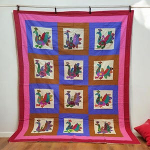 Könnte beinhalten: Eine Patchwork-Decke mit rosa Rand und einem blauen und braunen Gittermuster. Jedes Quadrat zeigt ein farbenfrohes gesticktes Pfauenmotiv.