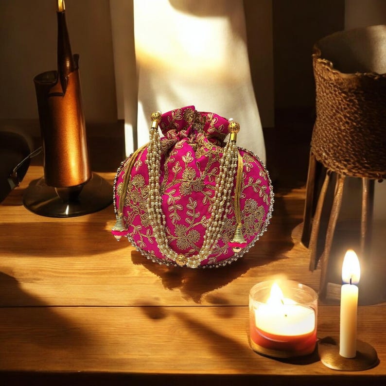 May include: A pink and gold embroidered drawstring bag with a pearl and gold tassel. The bag is round and has a floral design.
