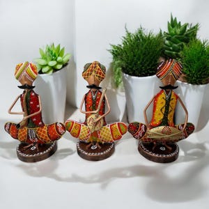 Puede incluir: Tres figuritas de metal coloridas de músicos tocando instrumentos tradicionales indios. Cada figurita lleva un turbante y un atuendo colorido. Las figuritas están sentadas sobre una base marrón.