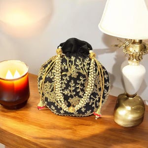 May include: A black drawstring bag with gold embroidery and pearl accents. The bag has a round shape and is decorated with a floral pattern.