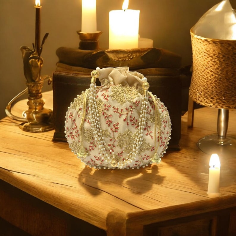 May include: A white and gold embroidered drawstring bag with a pearl and gold tassel. The bag is sitting on a wooden table with candles in the background.