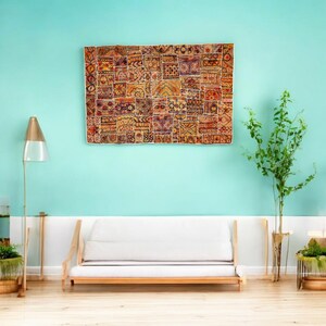 Op de afbeelding: Een groot, rechthoekig patchwork textiel met diverse geometrische en bloemmotieven in tinten rood, oranje, geel en bruin hangt aan een turquoise muur. Daaronder staat een witte bank met een houten frame. Een vloerlamp en een plant staan aan weerszijden.