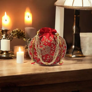 May include: A red, round, embroidered clutch bag with gold and white embellishments. The bag has a drawstring closure and a beaded tassel.